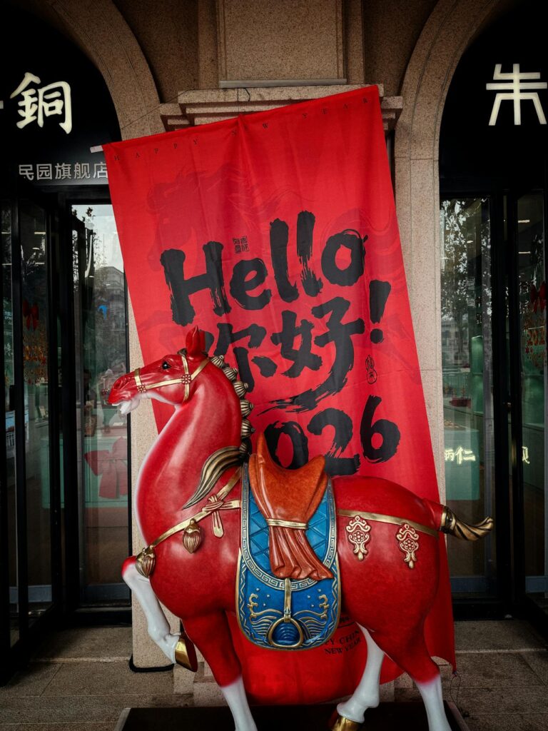 La foto raffigura una statua di piccole dimensioni di un cavallo rosso situata all'esterno di un negozio in Cina. Dietro di essa c'è un banner rosso con la scritta hello nihao 2026.