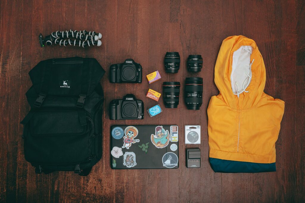 must have: zaino da viaggio con accanto un pc, delle macchine fotografiche, degli obbiettivi e una felpa gialla piegata