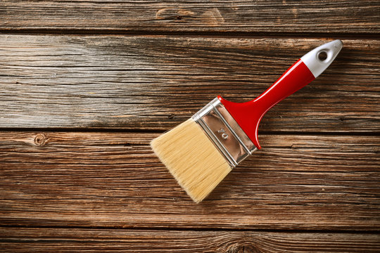 Un pennello da pittura con setole chiare, ghiera argentata e manico rosso e bianco è appoggiato su una superficie di legno scuro a listoni orizzontali. Il pennello è leggermente inclinato e occupa la parte superiore destra dell'immagine, lasciando spazio al legno sulla sinistra.