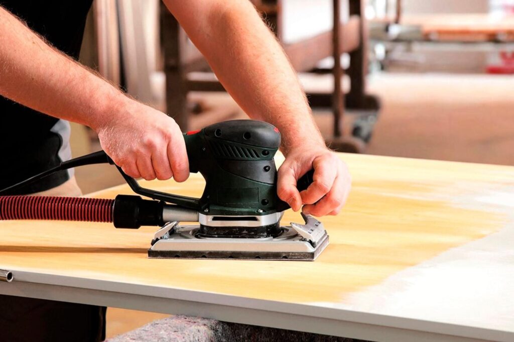 Due mani maschili stanno utilizzando una levigatrice orbitale nera e verde su una superficie di legno chiara. La levigatrice è collegata a un tubo rosso per l'aspirazione della polvere. Si nota la differenza tra la parte del legno già levigata, che appare più chiara e liscia, e la parte ancora grezza. Guida pratica in tre passaggi: come restaurare un vecchio tavolo.