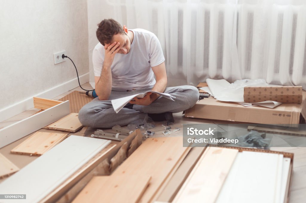 Uomo confuso mentre assembla mobili in legno. Difficoltà e svantaggi del montaggio fai-da-te dei mobili a casa senza l'aiuto di un esperto. L’uomo legge le istruzioni e non capisce cosa fare dopo.