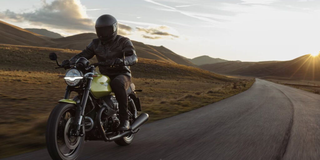 Pilota su Moto Guzzi V7 in assetto racing percorre una strada panoramica al tramonto.