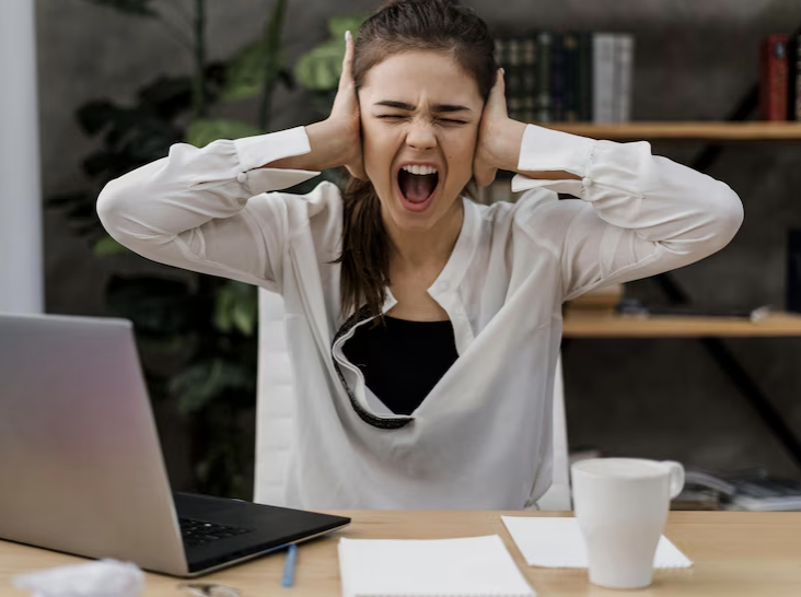 Donna che grida seduta sulla sua scrivania da lavoro con il computer davanti e dei documenti. Ha bisogno di consigli utili per uscire dal burnout.